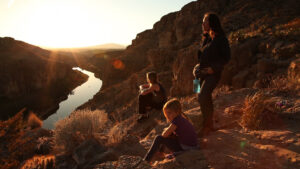An adult woman and two teens rest on rocks overlooking a river as the sun sets; a scene with Lulu Adams, Zelda Adams, and Toby Poser from the film Rumblestrips.